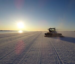 Photo looking down the runway with the snow grooming machine driving away as it is working the ice surface on a blue sky sunny day.