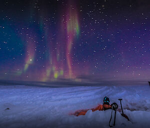 Expeditioner laying in snow during survival training, with an aurora lighting up the night sky