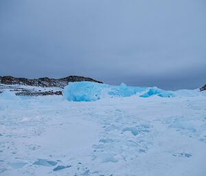 Blue iceberg with snow surrounding it and hills in the background