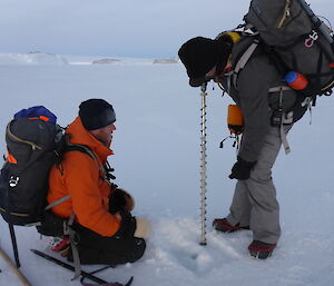 Expeditioner drilling ice with another looking on.