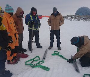 Expeditioners watching Mucles kneeling on snow, setting up for Hägg recovery briefing.