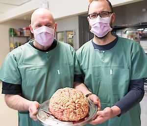 Andrew and Clint in scrubs with a cake that looks like a brain on a plate.