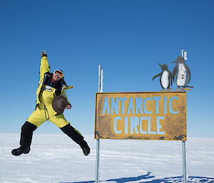 Expeditioner jumping for joy beside Antarctic Circle sign