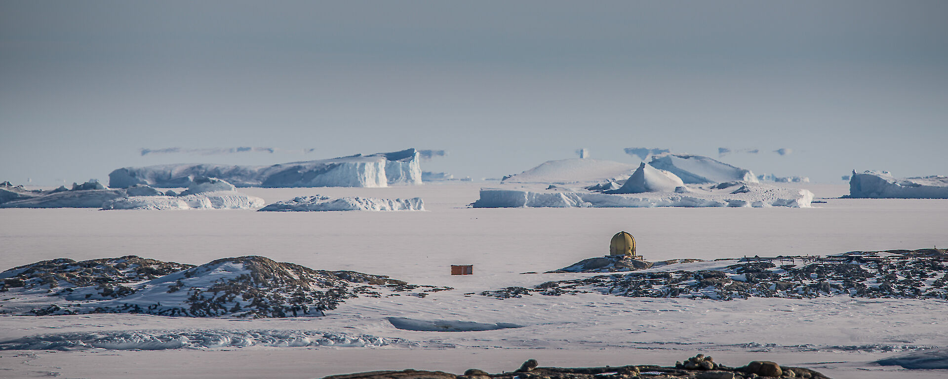 A landscape with sea ice at front, old station weather dome in mid shot and icebergs caught in the sea ice at top. Blue skies.