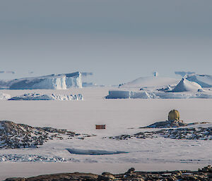 A landscape with sea ice at front, old station weather dome in mid shot and icebergs caught in the sea ice at top. Blue skies.