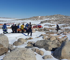 Snow and rocky foreground, Group of expeditioners undertaking a stretcher carry to a red Hägglunds waiting in the background.