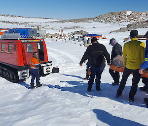 Group of expeditioners carrying a stretcher approaching the rear of a red hagglunds