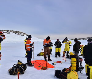 Group of expeditioners standing in circle on sea ice, Two in centre with drill bits preparing to drill a hole in the ice.