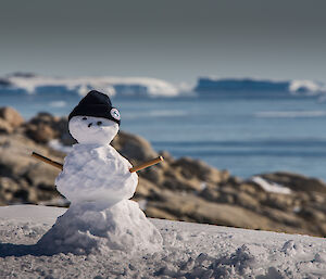 Snow man wearing a beanie in foreground with rocks in middle and icebergs in background.