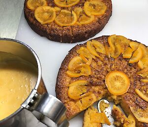 Two orange cakes, one at front right with piece cut out. Metal jug of custard at front left.