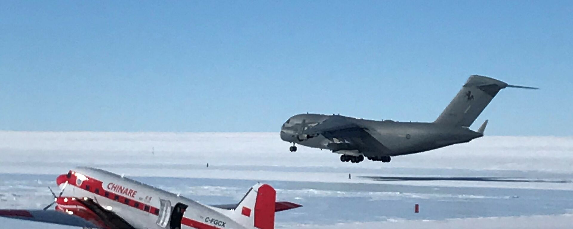 C-17, large grey military aircraft landing on ice runway with red and white basler aircraft on airfield apron in foreground.
