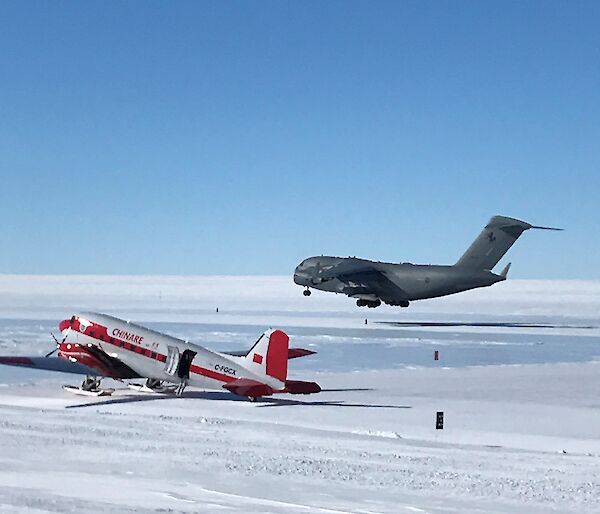 C-17, large grey military aircraft landing on ice runway with red and white basler aircraft on airfield apron in foreground.