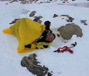 Expeditioner in yellow and black jacket with head and shoulders sticking out of yellow bivy bag which is lying on the snow mid picture
