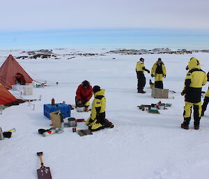 Two yellow and red pyramid shaped tents middle picture on left with crop of expeditioners in yellow and black gortex pants and jackets mid right and expeditioner on knees before camp stove in middle of picture. All standing on snow covered flat ground. Rocky horizen in background.