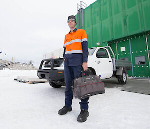 Expeditioner in high vis clothing carrying a tool box standing in front of a ute and large green building