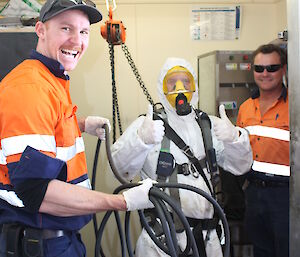 Person in white burn suit and breathing apperatus attached to winch giving the thumbs up, with two expeditioners in high vis either side.