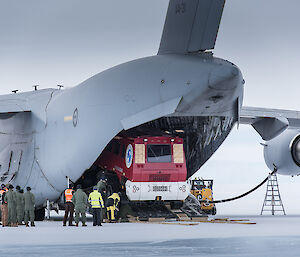 Large grey airforce plane with front of red bus seen coming out of rear cargo hold. Group of people in front of picture in kakhi airforce flying suits.