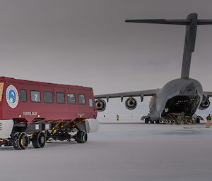 in foreground red bus on metal wheels with large grey airforce plane in background, all on blue ice runway