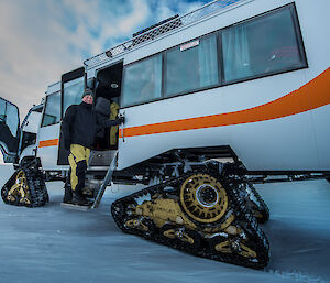 White bus with orange strip on tracked wheels, right foreground to left background. With man in yellow and black pants, black top and black beanie at door of bus.