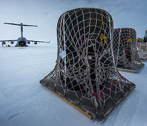 Four very large wheels on two pallets with netting over them in a line with large tractor in background. Left background is large grey airforce aircraft on ice runway.