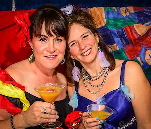Two ladies in bright formal dresses pictured from waist up holding cocktail glasses filled with an orange coloured drink.