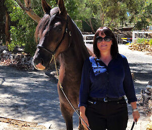 Lady in jeans, blue shirt and dark sunglasses, centre right, with chesnut horse standing beside her.