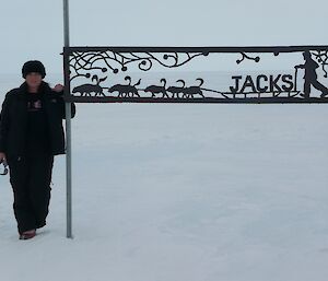 Lady in black overpants and jacket to left standing beside metal sign which has cut out of dogs sled with the word Jacks as the sled baggage.