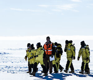 Man in black snow gear wearing orange hi vis vest, escorting a group of 6 school kids in yellow and black snow grear across snow covered ground