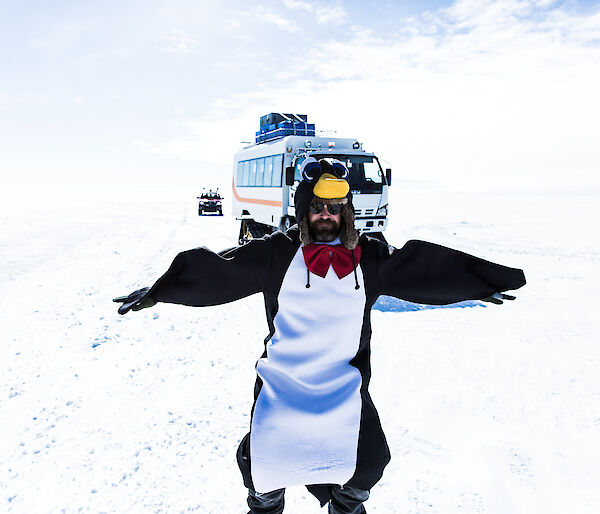 Man with beard and sun glasses dressed in penguin outfit in foreground with tracked white and orange bus behind.