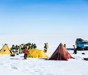 Two polar pryamid tents and one polar dome tent in middle of picture, large group of peoople in yellow and black gathered around tents with blue hagglands tracked oversnow vehicle to rear right of picture. All on white snow with blue sky.