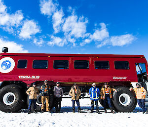 Large red bus with enormous wheels with group of seven expeditioners standing in front (heads just coming up to top of wheels). Bright blue sky behind.