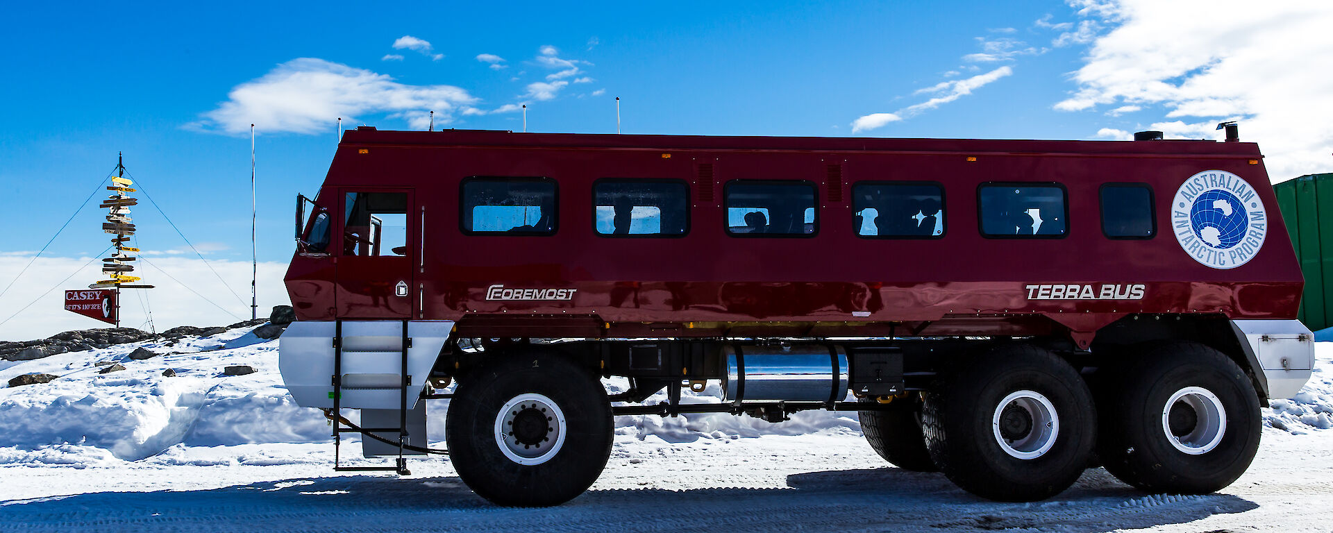 Large red bus with 1.5m high wheels, in foreground. In left background, casey sign with tags showing distance to Australian towns.