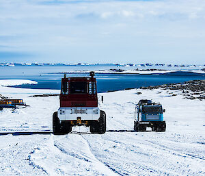 Snow covered slope in foreground, Blue water bay and blue sky in background. Centre picture, large red bus from back, with small blue hagglands travelling beside.