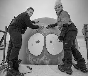 Top of orange melon with two painted eys with scaffolding at front with two expeditioners standing on with sandpaper.