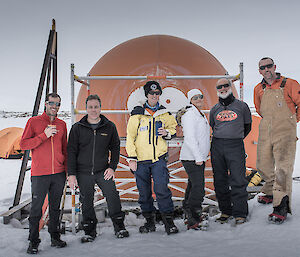 A group of six expeditioners standing in front of the orange melon hut.