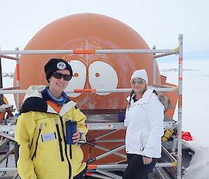 Orange melon hut with scaffolding in place, Two ladies in front, one in yellow jacket and black beanie to left and one in white jacket and beanie to right.