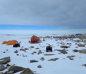 Grey sky, snow covered rocky ground in foreground. Mid picture orange melon hut with three yellow dome tents to left and two quad bikes to left and centre.