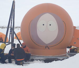 Large centre picture, orange melon hut with face painted onto it. Four expeditioners sitting to bottom left eating from plates.