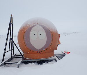 Orange melon dome with face painted, centre picture. Light dusting of snow over top of hut.