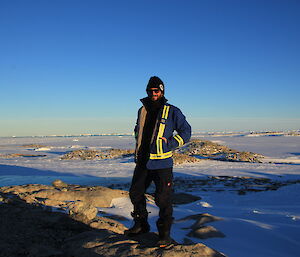 Man in beanie and sunglasses and big blue jacket with high vis trims standing on rock in centre frame, Background is ice and rocky islands with bright blue sky.