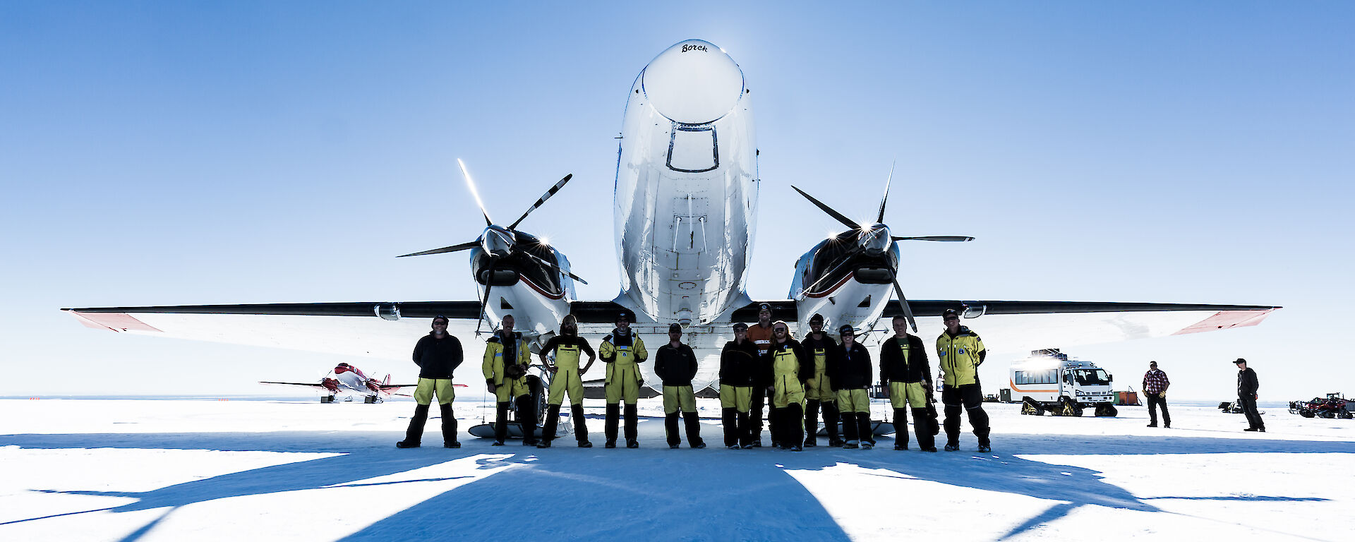 Group of expedtioners in yellow and black winter wear, standing in front of a twin propellored white plane which is parked on the ice. Blue sky above.