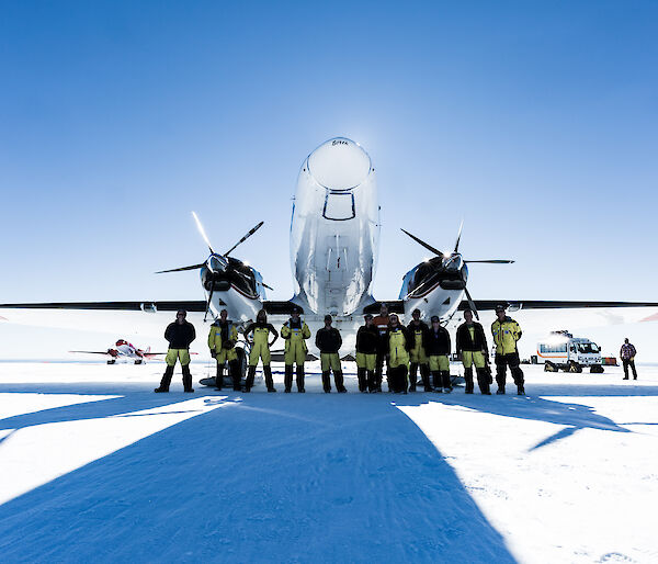 Group of expedtioners in yellow and black winter wear, standing in front of a twin propellored white plane which is parked on the ice. Blue sky above.