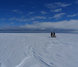 White snow covered ground to horizon, blue sky with white fluffy clouds covering approx 40% of sky. Mid frame, person on bike with person running beside.