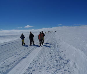 Snow covered ground with ploughed track to horizen, brigh blue ski above. Group of expeditioners running down the track in winter outfits.