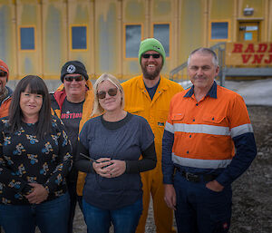 A group of six expeditioners in normal day wear, standing in front of a yellow building with sign in red writing saying ‘Radio VNJ'.