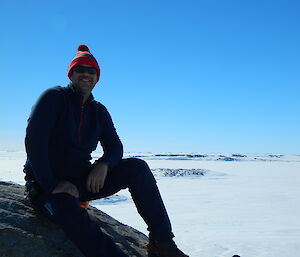 Curved rock in foreground with expeditioner in black pants and shirt and red beanie. Icy flat background to horizen (mid picture) with bright blue sky.
