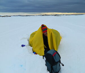 Snow covered ground, grey sky, expeditioner in stripey beanie in yellow bag with blue pack at front of bag