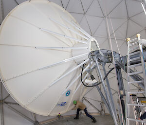 Inside white dome, with large white satellite dish in centre and left of picture. Man in brown jacket and green beanie pretending to push the satellite dish into position.