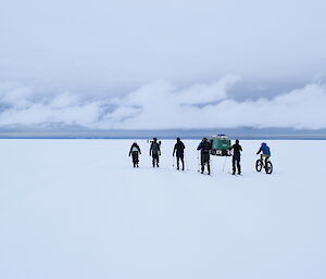 Snow covered ground to horizen(mid frame), grey sky with white band of cloud just above. Group of six expeditioners, mid frame, five skiing one on a bike, with green hagglands just in front.
