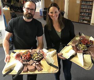 Man and woman both dressed in black, holding two very large platters covered in assorted cheese and meats.